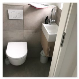 Der Raum zeigt eine minimalistische Toilette und ein Waschbecken, umgeben von grauen Fliesen und einem hellen Licht.