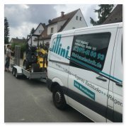 Ein Transporter mit einem Anhänger, auf dem eine gelbe Maschine steht, parkt an einer Straße in einem Wohngebiet.