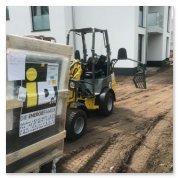 Ein gelber Minibagger steht auf einer Baustelle, umgeben von frischem Erdreich und einem Holzgestell mit einem Hinweis.