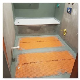Der Raum zeigt eine unfertige Badezimmerinstallation mit einer Badewanne und orangefarbenen Heizmatten auf dem Boden.