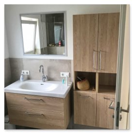 Der Waschtisch aus Holz hat eine weiße Arbeitsplatte und ist mit einem Wasserhahn sowie Pflegeprodukten ausgestattet.