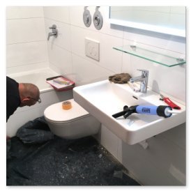 Im Badezimmer sind Werkzeuge und Materialien auf dem Boden verteilt, während ein Mann an der Wand arbeitet.